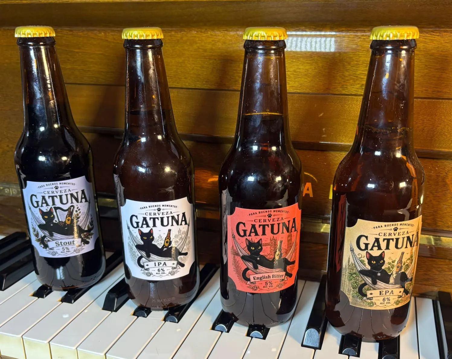 Las cuatro Cerveza Gatuna —Stout, IPA, English Bitter y EPA— apoyadas sobre las teclas de un piano de madera, con la tapa dorada brillando.